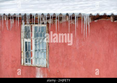 Eiszapfen hängen vom Dach über dem verbarrikierten Fenster eines alten Hauses mit einer roten Wand. Stockfoto