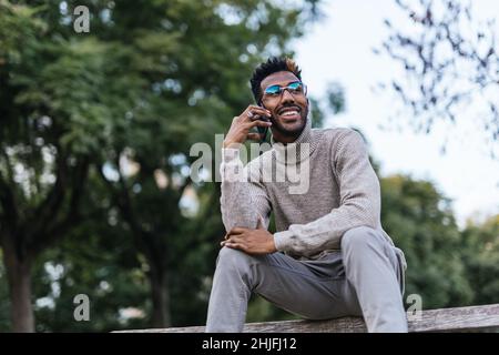 Lächelnder afroamerikanischer Mann in Rollkragenweste sitzt auf einer Parkbank und redet auf seinem Handy Stockfoto