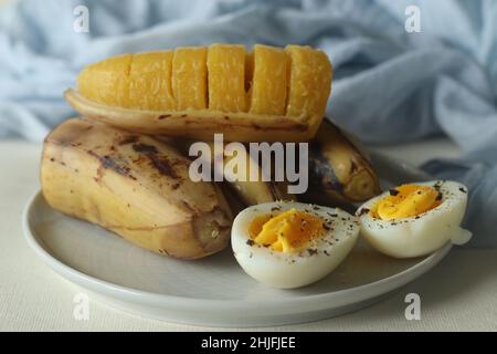 Gedämpfte Kochbanane und halbgeschnittenes gekochtes Ei mit zerdrücktem Pfeffer darauf. Aufgenommen auf weißem Hintergrund Stockfoto