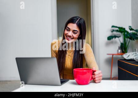 Porträt einer fröhlichen, fröhlichen jungen Frau, die zu Hause eine Tasse Kaffee genießt, während sie ein lächelndes Mädchen mit einem Laptop benutzt, das Tee oder heiße Schokolade trinkt, während sie Onli zubereitet Stockfoto