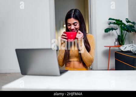 Glücklich zufrieden junge Frau genießen Tasse Kaffee zu Hause, während mit Laptop lächelndes Mädchen trinken Tee oder heiße Schokolade, während Online-Video cal Stockfoto