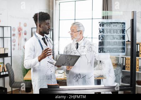 Multikulturelle männliche Ärzte stehen in der Nähe eines transparenten Flipchart mit Röntgenscan darauf. Zwei medizinische Mitarbeiter in Laborkittel schauten sich gegenseitig an, sprachen und benutzten einen modernen Laptop. Stockfoto