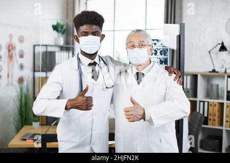 Junge afroamerikanische und alte kaukasische Ärzte in Gesichtsmasken und Laborkittel posieren in Innenräumen vor der Kamera. Zwei medizinische Spezialisten standen neben dem Flipchart und zeigten den Daumen nach oben Stockfoto
