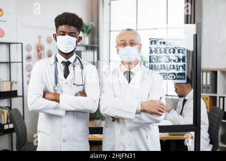 Junge afroamerikanische und alte kaukasische Ärzte in Gesichtsmasken und Laborkittel posieren in Innenräumen vor der Kamera. Zwei medizinische Spezialisten standen in der Nähe des Flipchart und hielten die Arme gekreuzt. Stockfoto