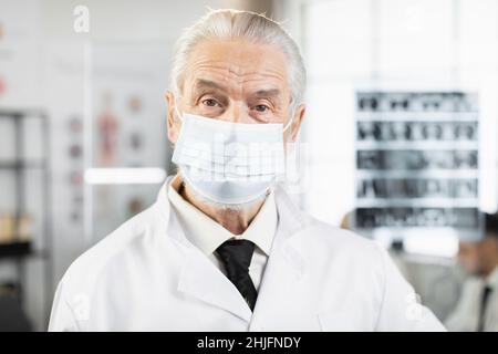 Qualifizierter Arzt im Alter mit Gesichtsmaske, Laborkittel und Brillen, die im Krankenhauszimmer stehen und die Kamera betrachten. Kaukasischer Arzt, der während des Arbeitsprozesses posiert. Stockfoto