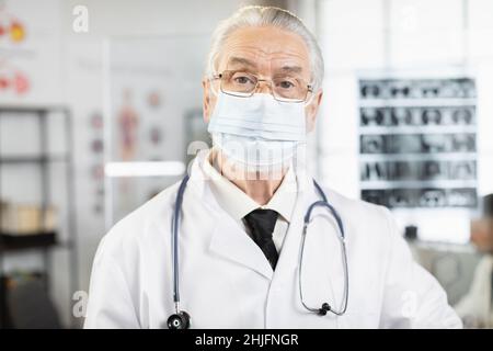 Qualifizierter Arzt im Alter mit Gesichtsmaske, Laborkittel und Brillen, die im Krankenhauszimmer stehen und die Kamera betrachten. Kaukasischer Arzt, der während des Arbeitsprozesses posiert. Stockfoto