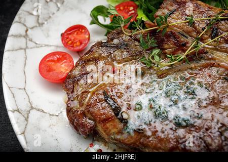 Gegrilltes Fleischsteak mit Butter, Kräutern und Kirschtomaten auf einem weißen Teller, vor dunklem Hintergrund Stockfoto