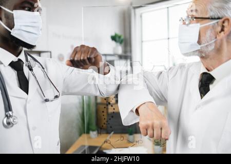 Zwei multirassische Gesundheitsexperten in Gesichtsmasken und Laborkittel begrüßen sich gegenseitig mit Ellenbogenanstrichen. Vorbeugende Maßnahmen während einer Coronavirus-Pandemie. Stockfoto