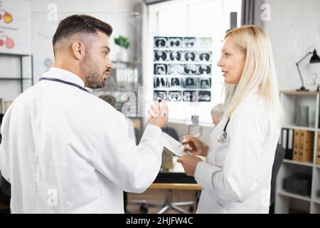 Zwei qualifizierte Ärzte in Laborkittel, die in der Nähe von Glasflipchart stehen und über die Ergebnisse der mrt sprechen. Kollegen bei der Besprechung im Konferenzraum. Diagnose und Krankheitskonzept. Stockfoto