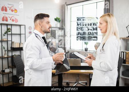Zwei qualifizierte Ärzte in Laborkittel, die in der Nähe von Glasflipchart stehen und über die Ergebnisse der mrt sprechen. Kollegen bei der Besprechung im Konferenzraum. Diagnose und Krankheitskonzept. Stockfoto