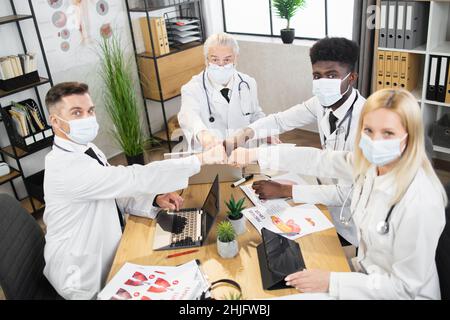 Eine Gruppe von multiethnischen Medizinern in schützenden Gesichtsmasken, die während des Treffens mit Fäusten zur Begrüßung anstoßen. Vier Ärzte, die mit modernen Geräten am Schreibtisch sitzen und die Kamera betrachten. Stockfoto