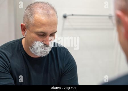 Ein hübscher weißhaariger bärtiger Mann, der sich seinen Bart abrasieren und in einen Spiegel schaut. Konzept eines sich ändernden Stils Stockfoto
