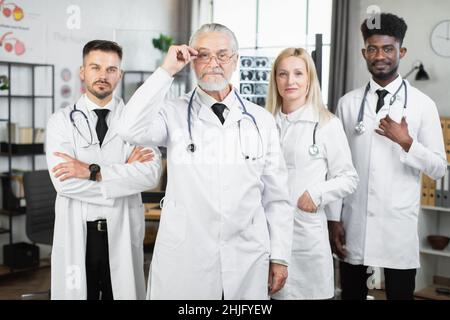 Leitender männlicher Arzt mit Augenmuschel und Laborkittel, der mit seinem multirassischen Team im Krankenhauszimmer steht und auf die Kamera schaut. Vier medizinische Mitarbeiter posieren nach dem Treffen in Innenräumen. Stockfoto