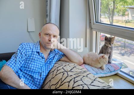 Porträt eines reifen Mannes und zweier niedlicher flauschiger Kätzchen auf einem Sofa am sonnigen Fenster Stockfoto