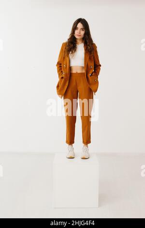 Junge Frau in modischem Pastellblazer und Hose mit stylischen Sneakers, die auf einem Würfel auf weißem Hintergrund im Studio steht. Stockfoto