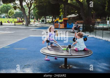 Zwei glückliche Schwestern spielen im Sommer auf dem Kinderspielplatz auf dem Karussell. Kinder drehen sich immer gerne schnell. Stockfoto