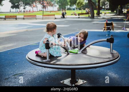 Zwei glückliche Schwestern spielen im Sommer auf dem Kinderspielplatz auf dem Karussell. Kinder drehen sich immer gerne schnell. Stockfoto