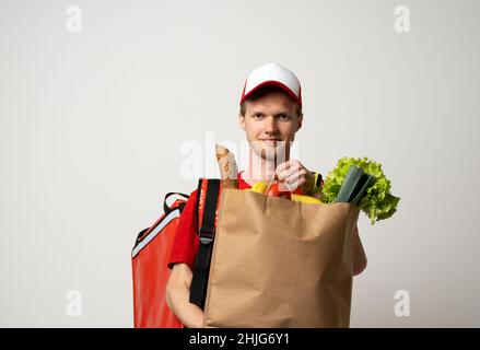 Happy Delivery Mann in roter Uniform mit Kappe und thermische Tasche Rucksack hält braune Bastelpapiertasche mit Lebensmitteln isoliert auf grauem Hintergrund Studio Stockfoto