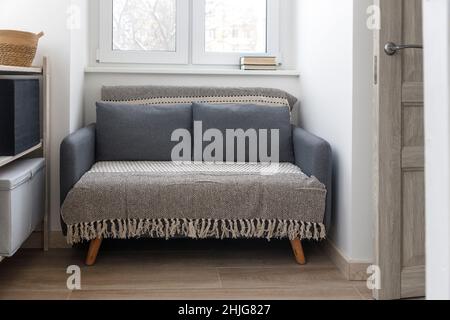 Graues weiches Sofa mit 2 Kissen. Modernes Design-Sofa im Stil von 70th in der Nähe des Fensters in einem kleinen leeren Raum. Speicherplatz kopieren. Stockfoto