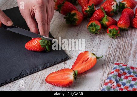Die Hände der Frau mit einem Messer schneiden Erdbeeren in zwei Hälften, auf einem alten weißen Holzhintergrund. Stockfoto