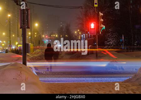 Im Winter steht ein Mann an einer Ampel, Autos mit Scheinwerfern fahren vorbei. Stockfoto