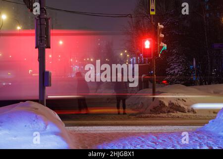 Im Winter steht eine Frau an einer Ampel, Autos mit Scheinwerfern fahren vorbei. Die rote Anzeige leuchtet. Stockfoto