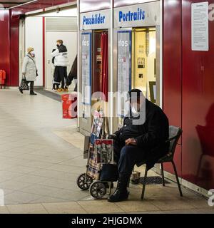 Ein Verkäufer der Straßenzeitung Biss sitzt mit Covid-Maske vor einer Fotokabine. Im Hintergrund ein junger Mann mit Maske umarmt seine Freundin Stockfoto