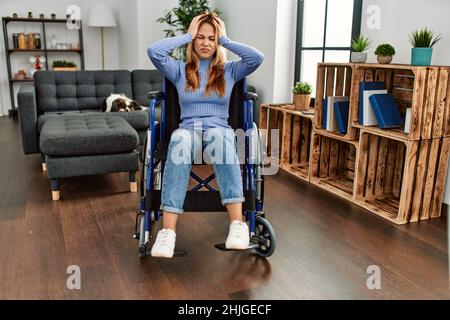 Junge schöne Frau, die zu Hause im Rollstuhl sitzt und unter Kopfschmerzen leidet, verzweifelt und gestresst, weil Schmerzen und Migräne. Hände am Kopf. Stockfoto