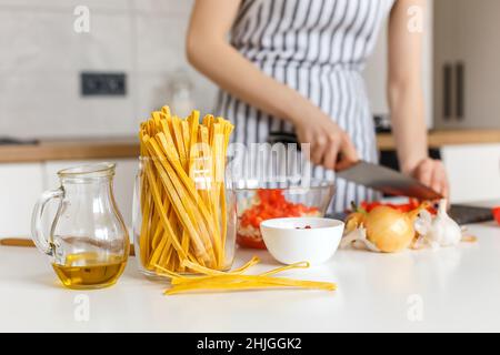 Zutaten aus der Nähe zum Kochen von Bolognese Pasta: Makkaroni, Tomaten, Olivenöl und Zwiebeln. Auf dem Hintergrund Frau, die traditionelle italienische Mahlzeit aus Nudeln zubereitet. Konzept der hausgemachten gesunden Lebensmitteln Rezept. Stockfoto