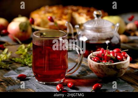 Hagebutten Beeren Kräutertee in Glas Becher stehen auf alten Holzbrett Tisch mit wilden Herbstbeeren und Apfelkuchen herum. Gesundes heißes Getränk. Dunkles rus Stockfoto