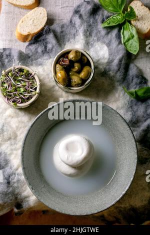 Traditioneller italienischer Burrata-Knüpfkäse in grauer Keramikschale auf dem Tisch. Brot, Oliven, grüner Salat. Zutaten für gesunde mediterrane din Stockfoto