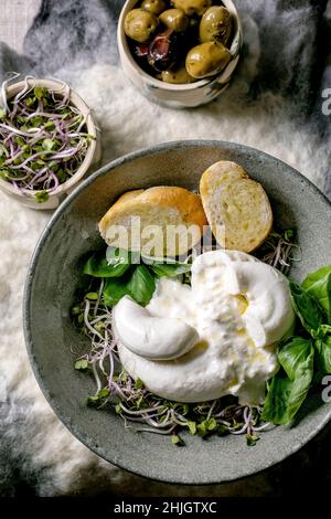 Traditioneller italienischer Burrata-Knüpfkäse-Salat in grauer Keramikschale auf dem Tisch. Brotscheiben, Oliven, grüne Sprossen. Gesundes mediterranes Dinn Stockfoto