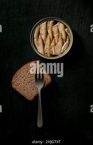 Offene Dose mit baltic Smoked Sprotten in Öl mit Roggenbrotscheibe und Gabel auf schwarzem Holzhintergrund. Draufsicht, Kopierbereich Stockfoto