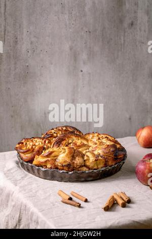Süße Apfel klebrige Zimtrollen Brötchen in runden Backblech stehen mit Gartenäpfeln und Zimtstangen auf grauem Leinentischtuch. Traditionell Stockfoto