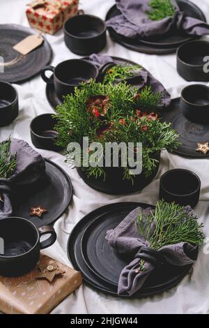 Weihnachtliche Tischdekoration mit leerem schwarzen Keramik-Geschirr, Tellern und Schalen, grünen Dekorationszweigen und Papierschachtel auf weißer Tischdecke Stockfoto