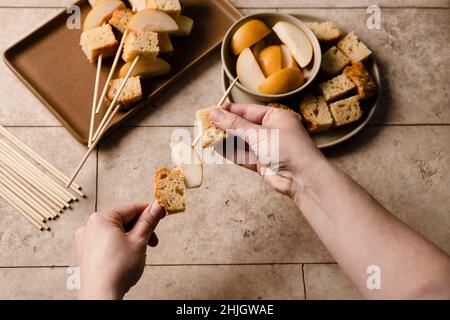 Hände, die Kuchenwürfel und Birnenscheiben auf einen Spieß legen. Stockfoto