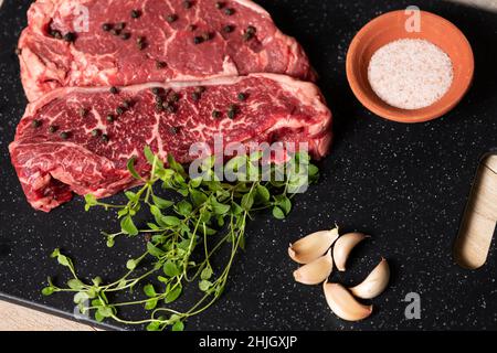 Zwei rohe, marmorierte Rindersteaks, fertig zum Kochen, mit frischem Olegano, Knoblauch und Pfefferkörnern. Stockfoto