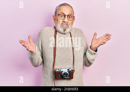 Gutaussehender älterer Mann mit Bart, der die Vintage-Kamera ohne Ahnung und verwirrt mit offenen Armen hält, keine Ahnung und zweifelhaftes Gesicht. Stockfoto