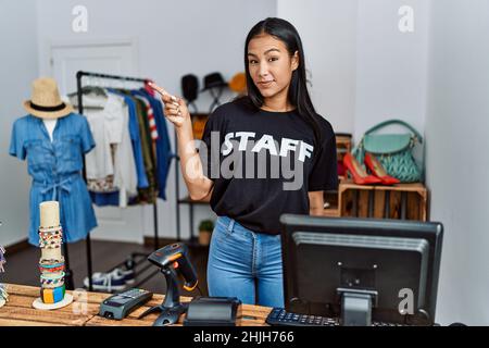 Junge hispanische Frau, die als Angestellte in der Einzelhandelsboutique arbeitet, zeigt mit dem Finger auf die Seite und zeigt Werbung, ernstes und ruhiges Gesicht Stockfoto