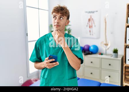 Junger kaukasischer Physio-Mann mit Smartphone in der medizinischen Klinik ernstes Gesicht denken über Frage mit Hand auf Kinn, nachdenklich über verwirrende Idee Stockfoto