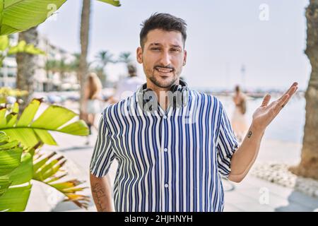 Junger, hübscher Mann, der im Freien mit Kopfhörern Musik hört, lächelt fröhlich und zeigt mit der Handfläche, die auf die Kamera schaut. Stockfoto