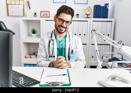 Junger Mann mit Bart in der Klinik mit Arztuniform und Stethoskop, die Hände zusammen und die gekreuzten Daumen lächeln entspannt und fröhlich. Erfolg Stockfoto