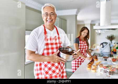 hispanische Paare mittleren Alters tragen Schürze Kochen hausgemachtes Gebäck sieht positiv und glücklich stehend und lächelt mit einem selbstbewussten Lächeln zeigt Zähne Stockfoto