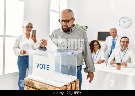 Wähler im mittleren Alter lächelnd glücklich Putting Stimme in der Wahlbox im Wahlzentrum. Stockfoto