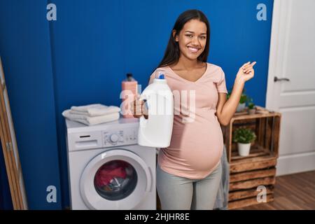 Junge Schwangerin, die Wäsche waschen und die Waschmittelflasche lächelnd hält, zeigt glücklich mit Hand und Finger zur Seite Stockfoto
