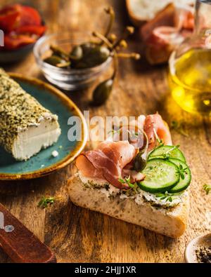 Sandwich mit hausgemachtem Prosciutto-Brot, Kräutern, Ziegenkäse, Auerkern, Gurken und Brunnenkresse auf Holzbrett. Antipasto. Bruschetta. Stockfoto