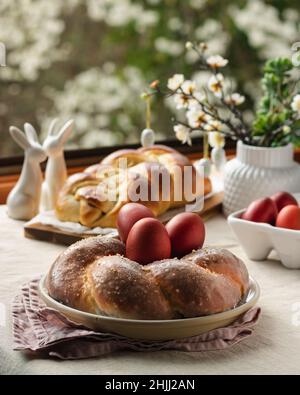 Osterbrot, österreichischer osternzopf, griechischer Tsoureki und rote Eier auf einem Tisch mit Leinentischdecke mit Blick auf das Frühlingsfenster, Stillleben Stockfoto