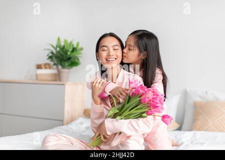 Lächelnd glücklich asiatische Teenager-Mädchen küssen und umarmen tausendjährige Frau mit Blumenstrauß im Schlafzimmer Stockfoto
