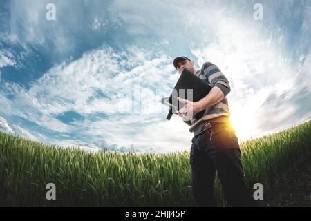 Ein junger Agronom hält eine Mappe in den Händen auf einem grünen Weizenfeld. Ein Landwirt notiert sich bei Sonnenuntergang über den Hintergrund landwirtschaftlicher Flächen. Mann Stockfoto