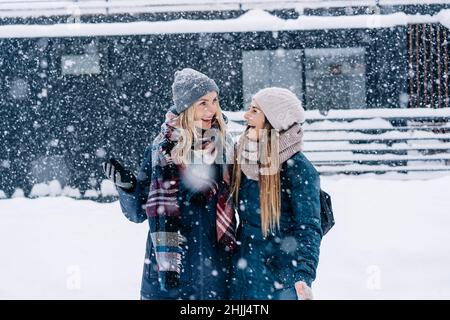 Zwei junge Frauen in warmer Winterkleidung stehen unter dem Schneefall und lachen fröhlich. Stockfoto